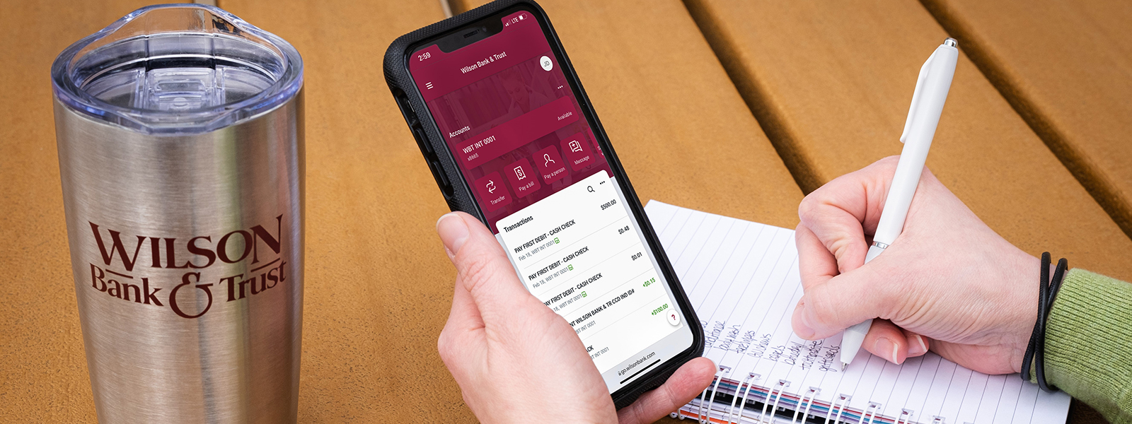 Someone holding a phone with their mobile banking app open and writing in a notebook.