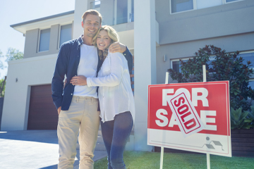 Young adult couple standing in front of two story house next to red "sold" sign