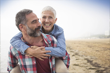 An older couple on the beach with the woman on the man's back smiling.