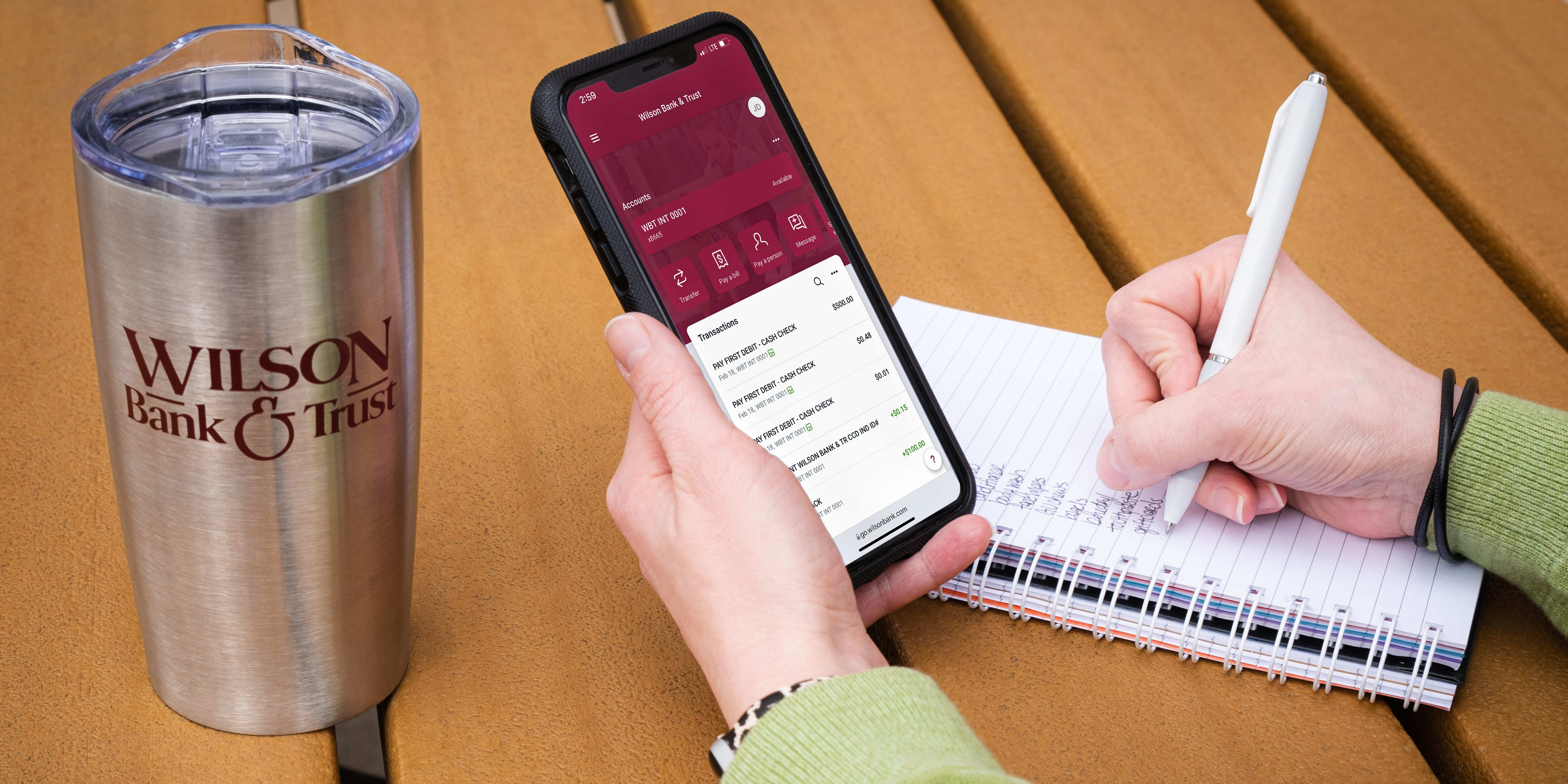 Someone holding a phone with their mobile banking app open and writing in a notebook.