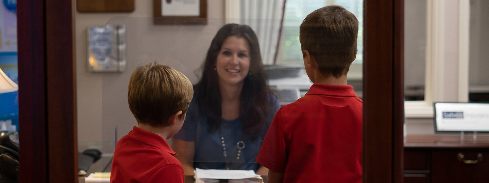 Two boys in red shirts stand in front of a woman's desk.