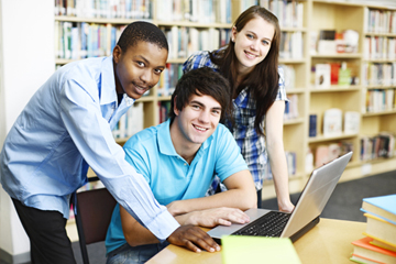 Two boys and a girl in a library smiling with a laptop in front of them.