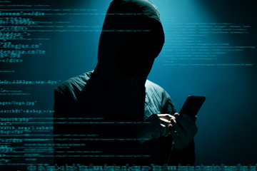 A person with a dark hood on holding a phone with code all around them.