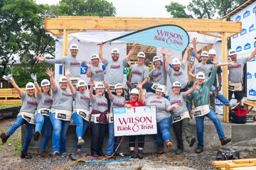 A group of people standing in front of a framing of a house with Wilson Bank & Trust shirts on.
